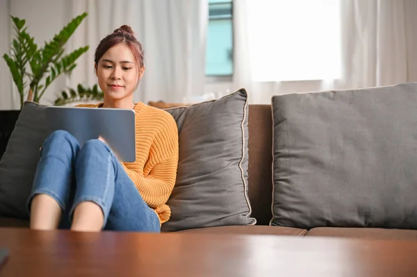 漂亮的年轻亚洲女人周末都呆在家里 坐在客厅舒适的沙发上 用便携式数码平板电脑 — 图库照片