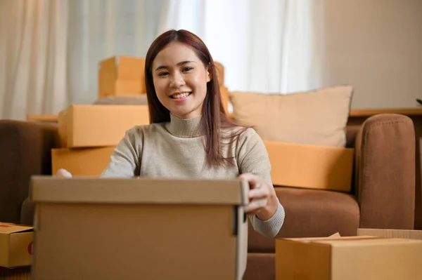 Happy millennial Asian woman packing her stuff in a cardboard box in the living room. House moving concept