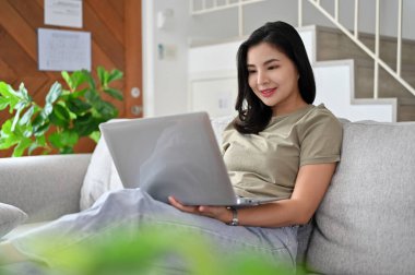 Yakışıklı genç Asyalı kadın, portatif dizüstü bilgisayarını kullanarak oturma odasındaki rahat kanepeye oturur, klavyeye yazar, internette sörf yapar..