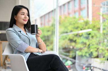 Kendinden emin ve başarılı Asyalı kadın yönetici ya da resmi giyimli iş kadını kahvesini yudumlayıp pencereden bakıyor, başarı tutkusu ve amacı hakkında hayal kuruyor..