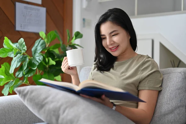 迷人迷人的年轻亚洲女人穿着休闲装 躺在舒适的沙发上 喝着早茶 在她的现代客厅里看书 — 图库照片