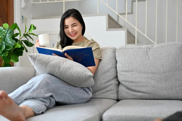 迷人的年轻亚洲女人在现代舒适的客厅里阅读一本关于自己闲暇时间的小说或教科书 — 图库照片