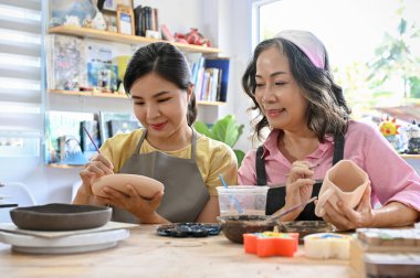 Mutlu ve neşeli Asyalı yaşlı kadın ve bin yıllık kadın seramik resim atölyesine katılıyor, stüdyoda seramik bir bardak çiziyor. Yaratıcı etkinlik kavramı