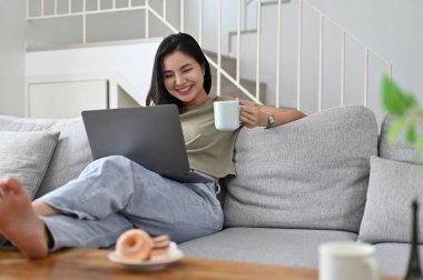 Mutlu ve neşeli Asyalı kadın hafta sonunu evde, kanepede kahve içerek ve dizüstü bilgisayar kullanarak geçiriyor..