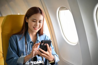 Güzel Asyalı bayan yolcu uçuş sırasında akıllı telefonunu kullanarak pencere kenarında oturur..