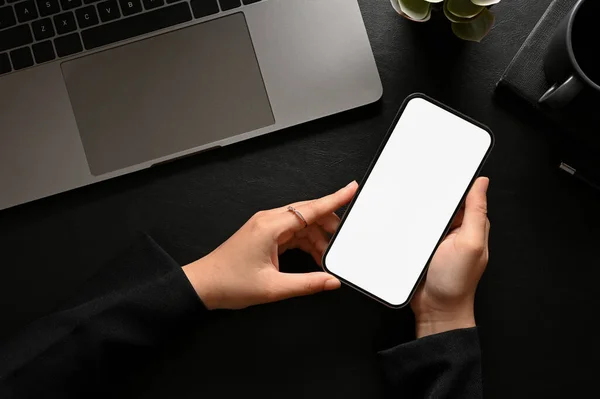 A female office employee or freelancer hands holding a smartphone white screen mockup over her modern dark and black hipster office desk. top view