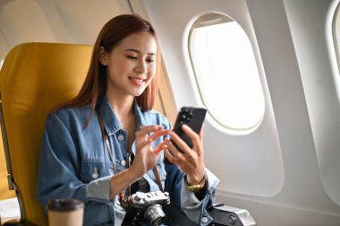 Güzel ve çekici Asyalı genç bir kadın uçuş sırasında akıllı cep telefonu kullanarak ekonomi sınıfında pencere kenarında oturuyor..