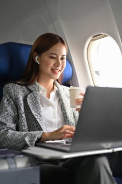 Uçakta resmi takım elbiseli çekici bir Asyalı iş kadınının portresi taşınabilir dizüstü bilgisayar kullanıyor ve uçuş sırasında sıcak bir kahve yudumluyor..