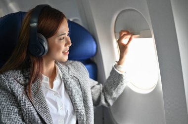 Güzel ve çekici Asyalı genç iş kadını ya da kablosuz kulaklıklı kadın pencere kenarına oturmuş güzel bir gökyüzü manzarası için pencereyi kaldırıyor..