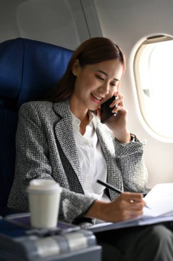 Meşgul ve çalışkan Asyalı bir iş kadını ya da iş ortağıyla telefonda konuşan, konferans için başka bir şehre uçarken kağıt üzerinde notlar alan bir kadın girişimci..