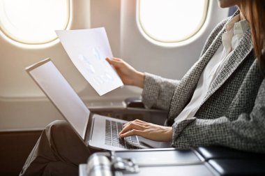 Profesyonel başarılı Asyalı iş kadını uçağa binip konferansa gidiyor, uzaktan kumandalı uçuş yapıyor, belge okuyor ve taşınabilir laptop kullanıyor. kırpılmış, dikiz görüntüsü
