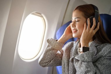 Güzelim ve güzel Asyalı iş kadını gündelik kıyafetleriyle uçuş sırasında modern kablosuz kulaklıklarıyla müzik dinliyor. Uçak taşımacılığı kavramı.