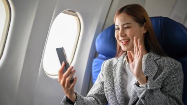 Resmi takım elbiseli mutlu ve neşeli Asyalı iş kadını akıllı telefon kullanıyor, başka bir şehre uçarken ailesiyle görüntülü görüşme yapıyor. Kentsel ulaşım kavramına sahip şehir yaşam tarzı.