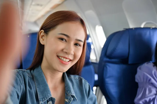 Beautiful young asian female travel blogger taking selfie or recording herself while she\'s sits at the middle seat in the economy class. Travel concept