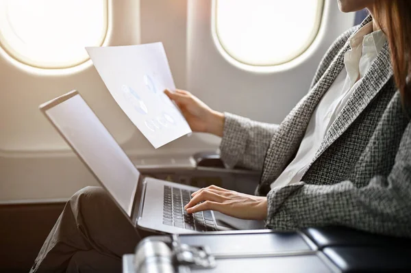 A professional successful asian businesswoman on a plane going to aboard meeting conference, remote working on a flight, reading a document and using portable laptop. cropped, rear view image