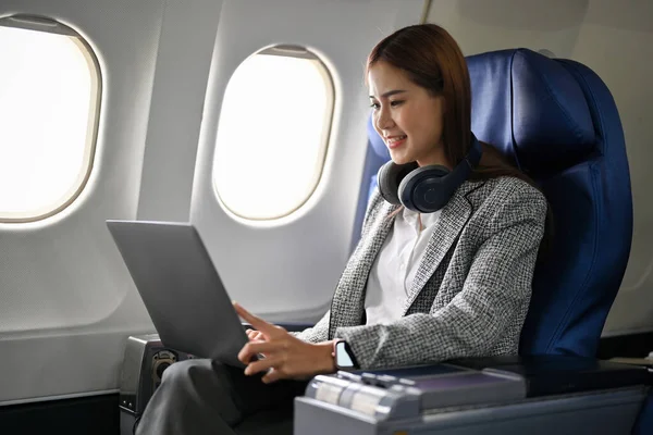 Beautiful and successful millennial asian businesswoman or female executive manager sits at her business seat on the plane, using portable notebook laptop during the flight.