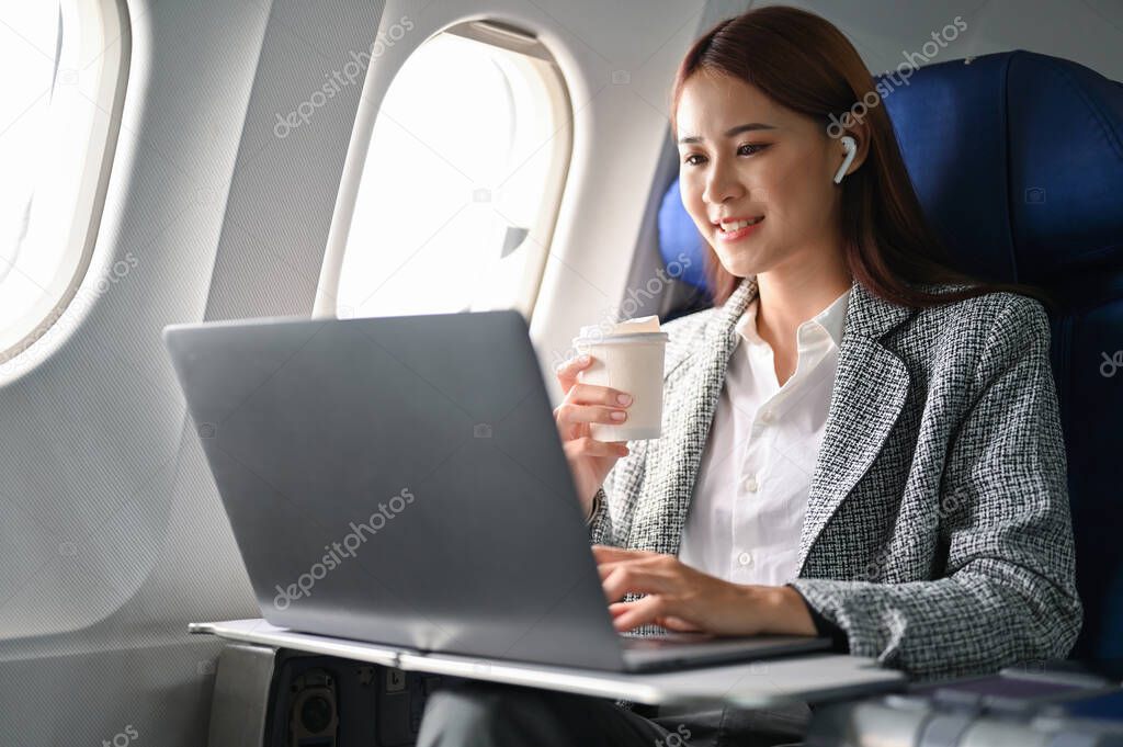 Successful and confident millennial asian businesswoman in formal suit on the plane using portable notebook laptop and sipping a hot coffee during flight.