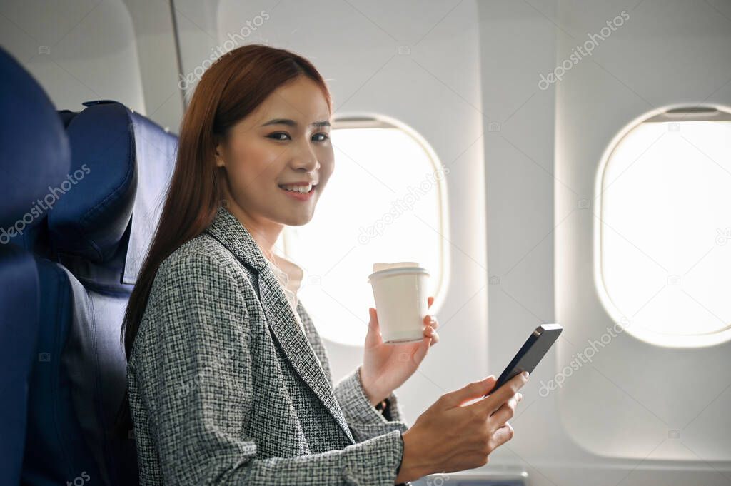 Charming and attractive millennial asian businesswoman or female entrepreneur using smartphone and sipping a hot coffee during flying to somewhere by plane.