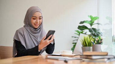 Güzel Müslüman iş kadını ya da kadın yönetici tesettüre oturmuş akıllı telefon kullanıyor..