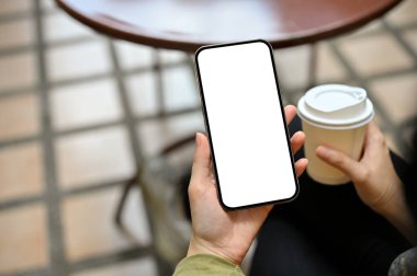 Bir dişi, güne başlamak için açık havada bir kafede akıllı telefon kullanıyor ve sabah kahvesini yudumluyor. Cep telefonu beyaz ekran modeli.