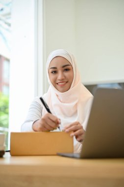 Çekici Asyalı Müslüman kadın küçük bir e-ticaret girişimcisi bir kargo kutusu paketliyor, karton bir kutuya müşteri adresi yazıyor..