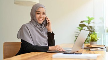 Laptop bilgisayarı projesinde çalışırken tedarikçisiyle telefonda tesettürlü ya da başörtüsü takan başarılı Asyalı Müslüman iş kadını.