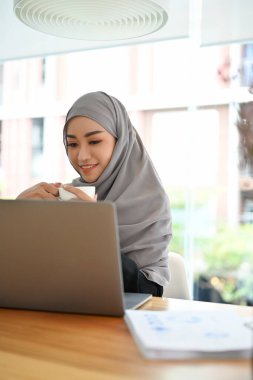 Başörtüsü ya da başörtüsü takan Asyalı genç ve güzel bir iş kadınının portresi ofis masasında dizüstü bilgisayarla oturuyor..