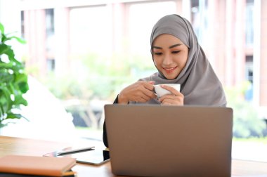 Güzel Müslüman iş kadını sabah kahvesini yudumluyor, laptop ekranına bakıyor, dizüstü bilgisayarından projesini kontrol ediyor..