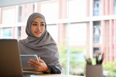 Çekici Müslüman iş kadını ya da tesettürlü kadın ofis masasında elinde dijital tabletle oturuyor..