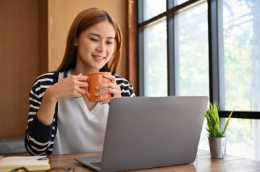 Sakin ve rahat bir Asyalı kadın sabah kahvesini yudumlarken masadaki taşınabilir bilgisayarından online içeriği okuyor..
