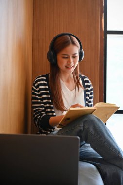 Çekici genç Asyalı üniversite öğrencisi kulaklıkla müzik dinliyor ve deftere bir şeyler yazıyor..