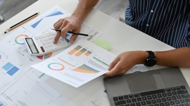 Masasında çalışan profesyonel bir erkek finansal yatırım yöneticisi, rapordaki finansal ve muhasebe satışlarını analiz ediyor, hesap makinesi kullanıyor..