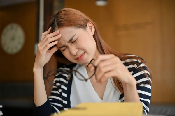 Stressed young Asian female college student or freelancer working at her desk, suffering from her headache or migraine symptom.