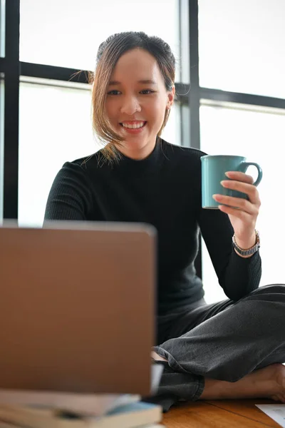 Beautiful young Asian female freelancer or female journalist working on her assignment on laptop computer and sipping morning coffee.