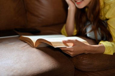 Relaxed young Asian woman laying on comfy sofa and reading a book or novel in her living room. cropped image