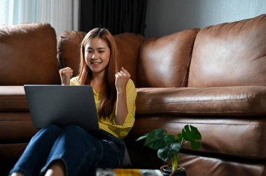Excited and pretty millennial asian woman in the living room using laptop computer to check her job offer email.