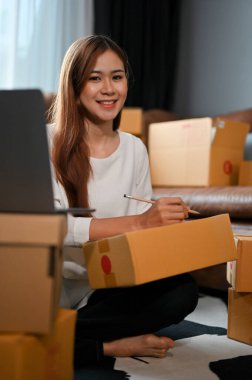 Çekici genç Asyalı bayan küçük online işletme sahibi oturma odasında nakliye kutuları hazırlıyor, müşteri paketini paketliyor, karton kutuya bir müşterinin adresini yazıyor..