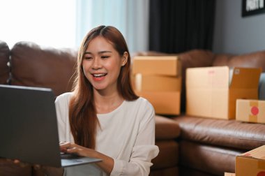 Happy and cheerful young Asian female SME online small business shop owner checking her sales orders on the website via laptop computer.