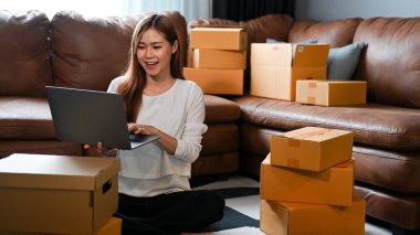 Beautiful and successful millennial asian female online shop owner replying a customer or checking stock on laptop computer in the living room. E-commerce business concept