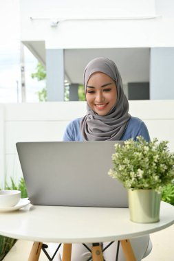 Attractive young Asian Muslim woman with headscarf or hijab remote working at home, using laptop computer.