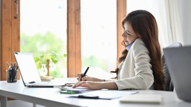 Attractive asian businesswoman or female secretary talking on the phone with business client at her office desk.