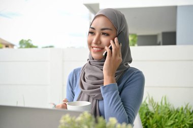 Beautiful and elegant millennial Asian Muslim woman wearing hijab talking on the phone with someone in house's backyard.