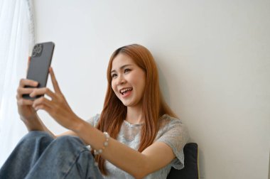 Güzel Asyalı kız oturma odasında rahatlamış, akıllı telefonunu kullanarak selfie çekiyor ya da kendi videosunu çekiyor..