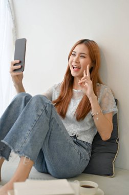 Güzel ve yakışıklı Asyalı genç bir kadın, oturma odasında modern akıllı telefonuyla selfie çekiyor..