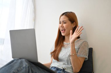 Cheerful and pretty young Asian woman having a video call with her friends on laptop computer at home.