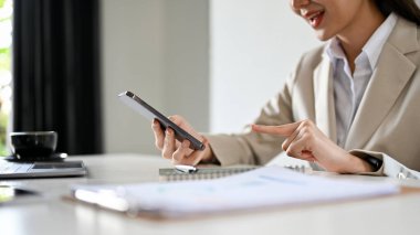 Çekici ve başarılı Asyalı bir bayan yönetici ya da resmi takım elbiseli bir iş kadını ofis masasında oturur ve biriyle mesajlaşmak için akıllı telefon kullanır. kırpılmış atış