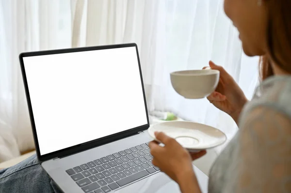 Close-up image, Beautiful young Asian female sipping a hot coffee while using laptop computer. Laptop white screen mockup.