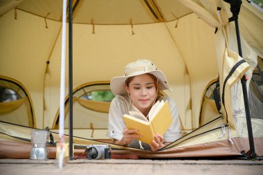 Rahat ve huzurlu genç Asyalı kadın kamp çadırında uzanıyor ve en sevdiği kitabı okuyor. Açık hava etkinliği kavramı.