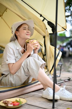 Rahat ve huzurlu genç Asyalı kampçı kadın çadırının önünde oturuyor, sabah kahvesini yudumluyor ve güzel doğa manzarasına bakıyor..
