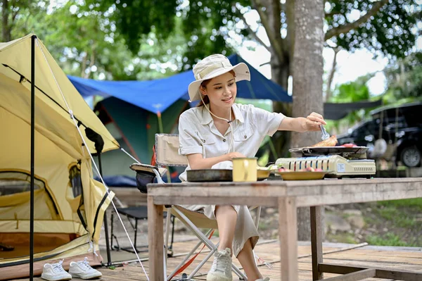 Attractive young Asian female camper grill some pork-chop steaks on a portable outdoor picnic stove, cooking some food with camping kitchen equipments at campground.